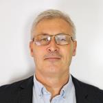 Portrettbilde av en glattbarbert mann med kort, grått hår og briller. Han har dressjakke og skjorte som er åpen i halsen.