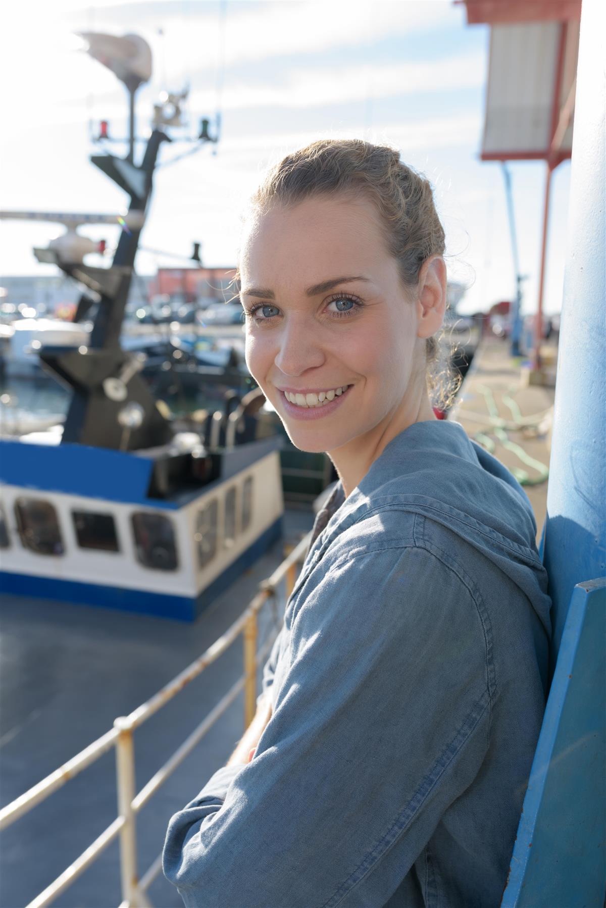 En ung kvinne står på dekket av en fiskebåt. Hun vender hodet mot kamera og smiler. Håret er bundet i en hestehale. - Klikk for stort bilde