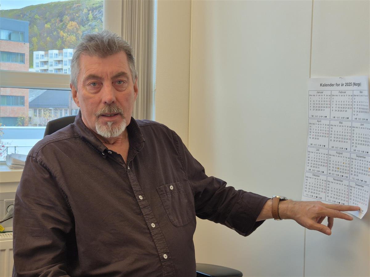 Mann med kortklipt, grått hår og skjegg sitter på et kontor foran vinduet og peker på en kalender. - Klikk for stort bilde