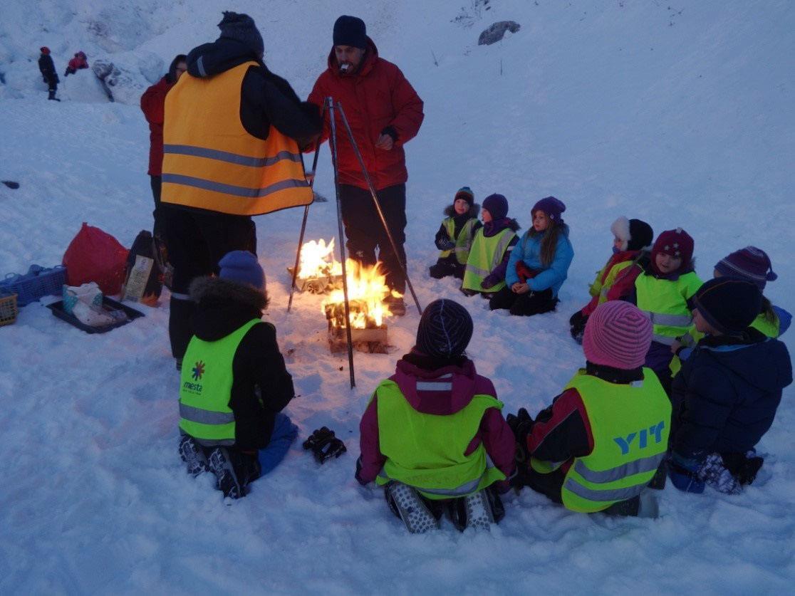 En gruppe barn med refleksvester sitter i ring rundt et bål i sneen. Tre voksne står ved bålet og ordner. - Klikk for stort bilde