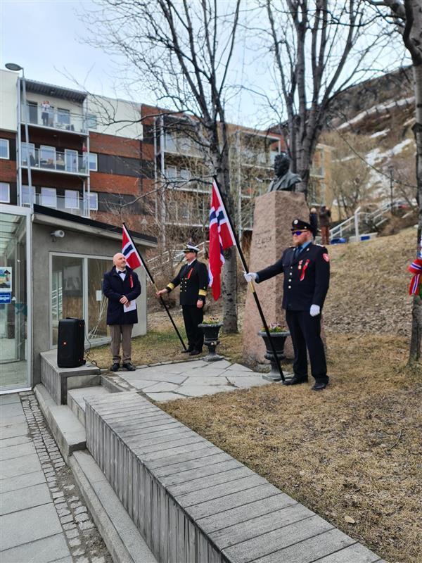 Ole Olsen statuen. To brannfolk i uniform holder norske flagg. Mann i dress holder tale. - Klikk for stort bilde
