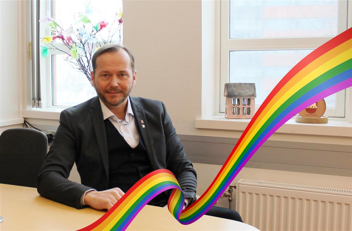 Portrettfoto av mann i dress uten slips ved et skrivebord. Et pridebånd i forgrunnen. - Klikk for stort bilde