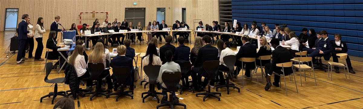 Elever sitter rundt et stort møtebord med skilt for ulike land under FN-rollespillet på Breilia skole - Klikk for stort bilde