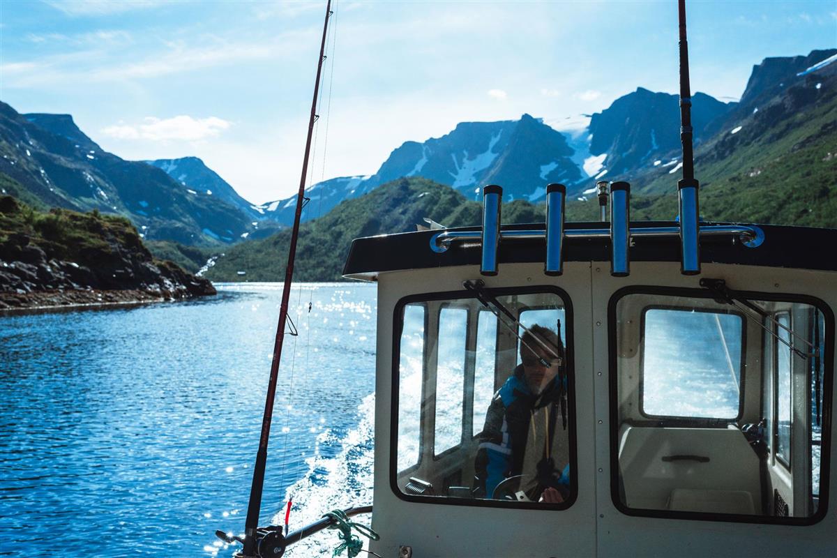 Mann står i styrhuset på liten båt som setter ut fjorden i solskinn - Klikk for stort bilde